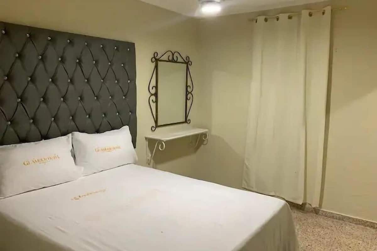 A hotel room with a tufted headboard, a bedside table with a mirror, and a bed with white bedding.