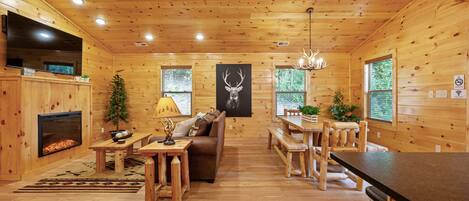A wooden cabin interior with a fireplace, a sofa, a dining table, and a chandelier.