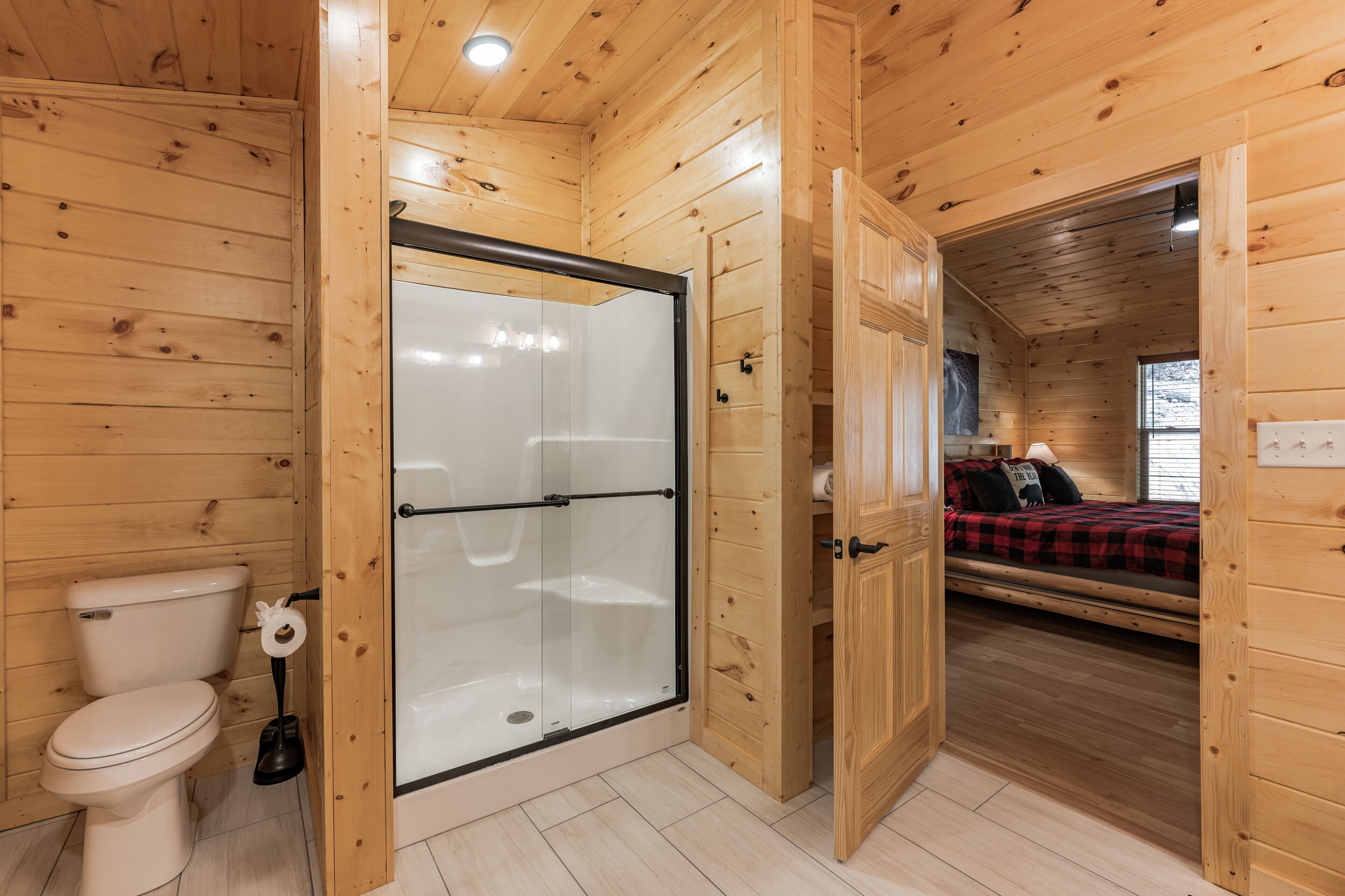 A wooden cabin interior with a glass-enclosed shower, a toilet, and a bed with a red and black checkered blanket.