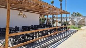 Een buitenruimte met een houten pergola, tafels en stoelen, omgeven door palmbomen en een stenen muur.