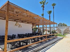 Area esterna con sedute, pergola in legno, tavoli e sedie, circondata da palme e un muro in pietra.