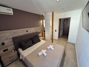 Ein modernes Hotelzimmer mit einem Holzkopfteil, einem Bett mit einer beigen Bettdecke, zwei Kissen, einem Nachttisch mit Telefon und Fernbedienung sowie einer Klimaanlage an der Wand.