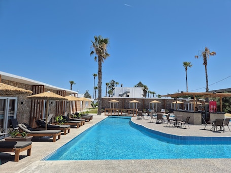 A pool area with lounge chairs and umbrellas, surrounded by palm trees and stone buildings.