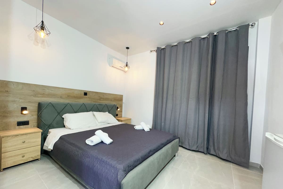 A modern hotel room with a bed, nightstands, a wooden headboard, and a large window with curtains.