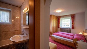 A hotel room with a bathroom, a bed, a bedside table, and a window with curtains. - 179A Apartments Seida (Selva di Val Gardena)