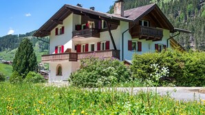 Un chalet de dos pisos con balcón, rodeado de exuberante vegetación y montañas al fondo.