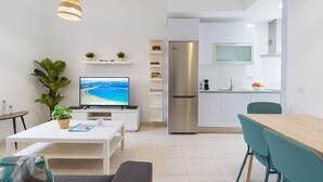 A modern living room with a flat-screen TV, a sofa, a dining table, and a kitchen area with a refrigerator and a microwave.