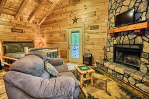 L’intérieur d’un chalet confortable, avec des murs en rondins de bois, une cheminée en pierre, un canapé confortable et un lit offrant une vue sur l’extérieur.