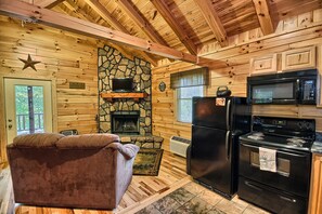 Interior acogedor de una cabaña con chimenea de piedra, televisor, refrigerador y zona de cocina.