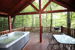 A wooden deck with a hot tub, picnic table, and chairs, surrounded by a forest.