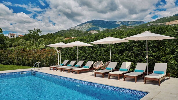 Una piscina con sillones de descanso y sombrillas, rodeada de vegetación y montañas.