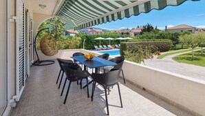 Un área de estar al aire libre cubierta con mesa y sillas, una piscina al fondo y vistas a casas y follaje.