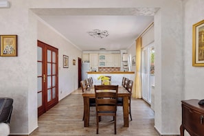 Un coin repas avec une table et des chaises en bois, une cuisine à l’arrière-plan et une vue à travers une porte vitrée.