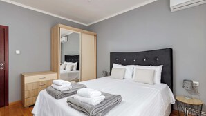 Una cama bien hecha con ropa de cama blanca y gris, una mesita de noche de madera con una lámpara, y un armario con espejo.