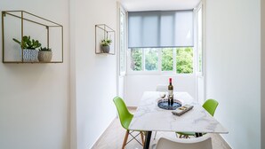 A dining area with a table, chairs, and a window with a blind. There are potted plants on a shelf and a wine bottle on the table.