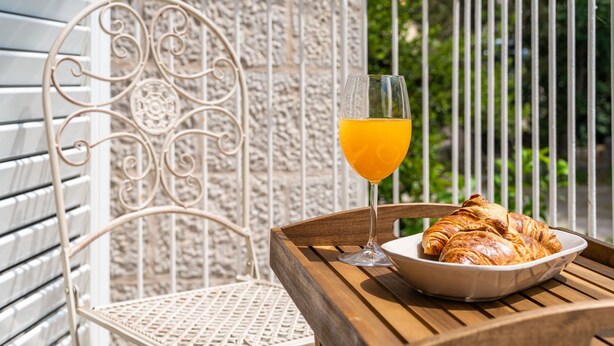 A wooden table with a bowl of croissants and a glass of orange juice on a balcony with a white metal chair and railing.