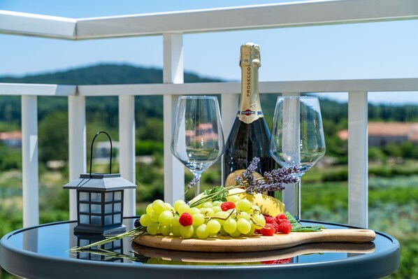 A table with a bottle of champagne, two glasses, a lantern, and a fruit platter on a balcony with a scenic view.