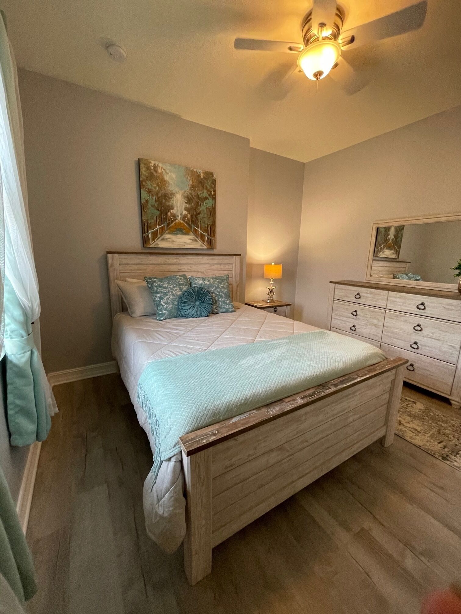 A bedroom with a wooden bed, a nightstand with a lamp, a dresser, a painting on the wall, and a ceiling fan.