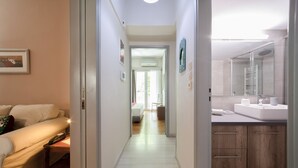 Ein Flur, der in ein Schlafzimmer mit Bett, Nachttisch und Stuhl führt. Es gibt ein Badezimmer mit Waschbecken, Spiegel und Handtuchhalter.