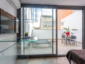 Eine moderne Wohnung mit einer Glastür zum Balkon. Innen befindet sich ein Glastisch mit Büchern, eine Bank und ein an der Wand befestigter Fernseher.
