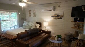 Une chambre à coucher comprenant un lit, un canapé en cuir marron, une table de chevet, une lampe, un appareil de climatisation, une télévision et une petite table ronde agrémentée d’une plante.