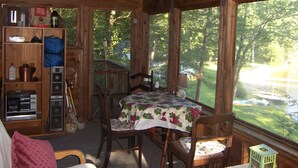 A rustic cabin interior with a dining table, chairs, a small table with a lamp, and a view of a lake through large windows. - Cozy Cabin on Loon Lake (Plymouth)