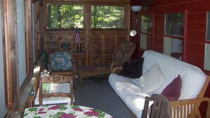 A cozy cabin interior with a wooden ceiling, a white couch, a floral tablecloth, and a view of the forest through large windows. - Cozy Cabin on Loon Lake (Plymouth)