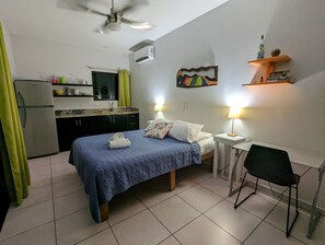 Habitació d'hotel senzilla amb llit, escriptori, cadira, petita cuineta i ventilador de sostre.