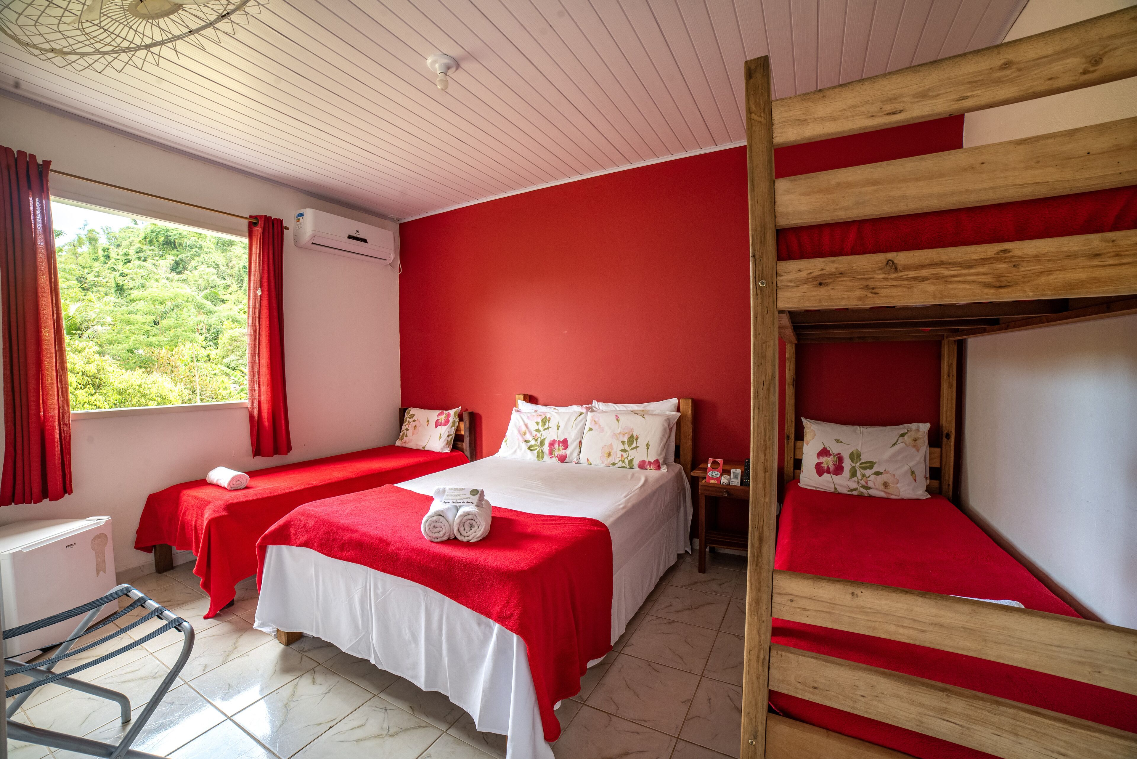 Quarto com duas camas, uma beliche, uma escrivaninha e uma janela com cortinas.