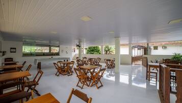 A spacious indoor area with multiple wooden tables and chairs arranged in clusters, large windows allowing natural light, and a high ceiling with recessed lighting.