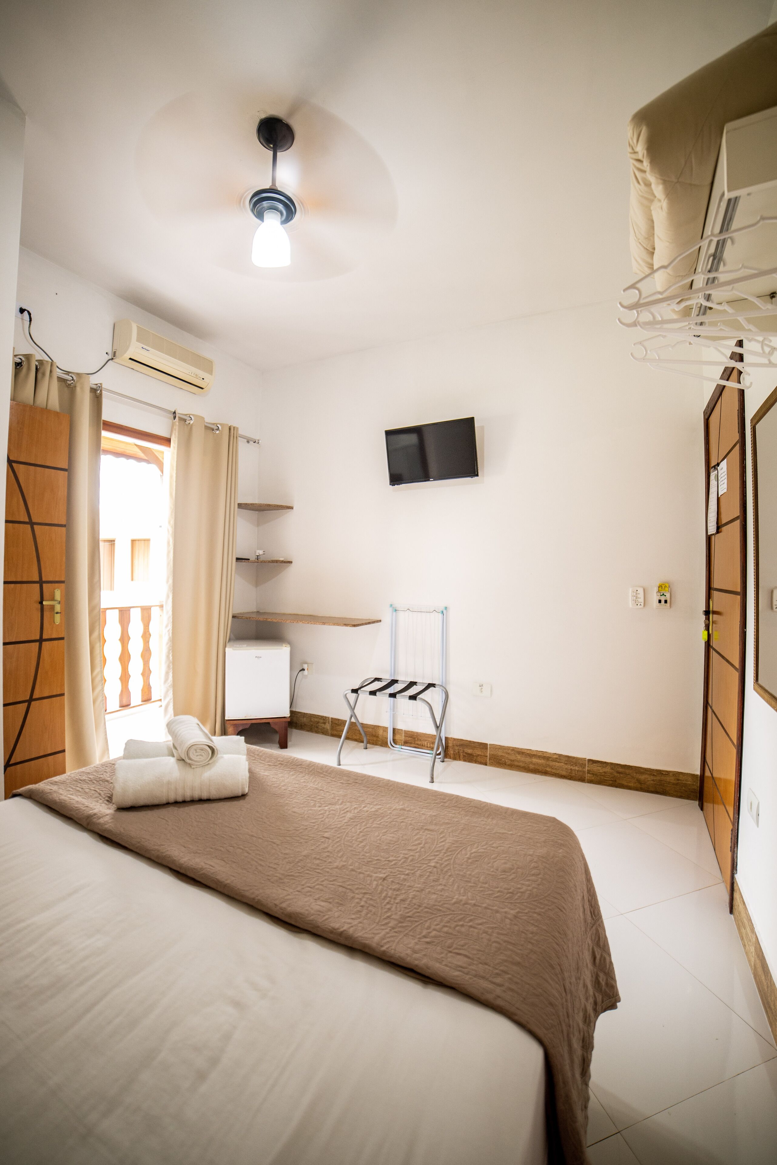 A hotel room with a bed, a ceiling fan, a TV mounted on the wall, an air conditioning unit, a wooden wardrobe, and a window with curtains.