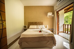 Une chambre d’hôtel dotée d’un grand lit, d’une tête de lit en bois, d’une table de chevet et d’une étagère murale où sont rangées des serviettes, ainsi que d’un balcon offrant une vue sur la verdure.