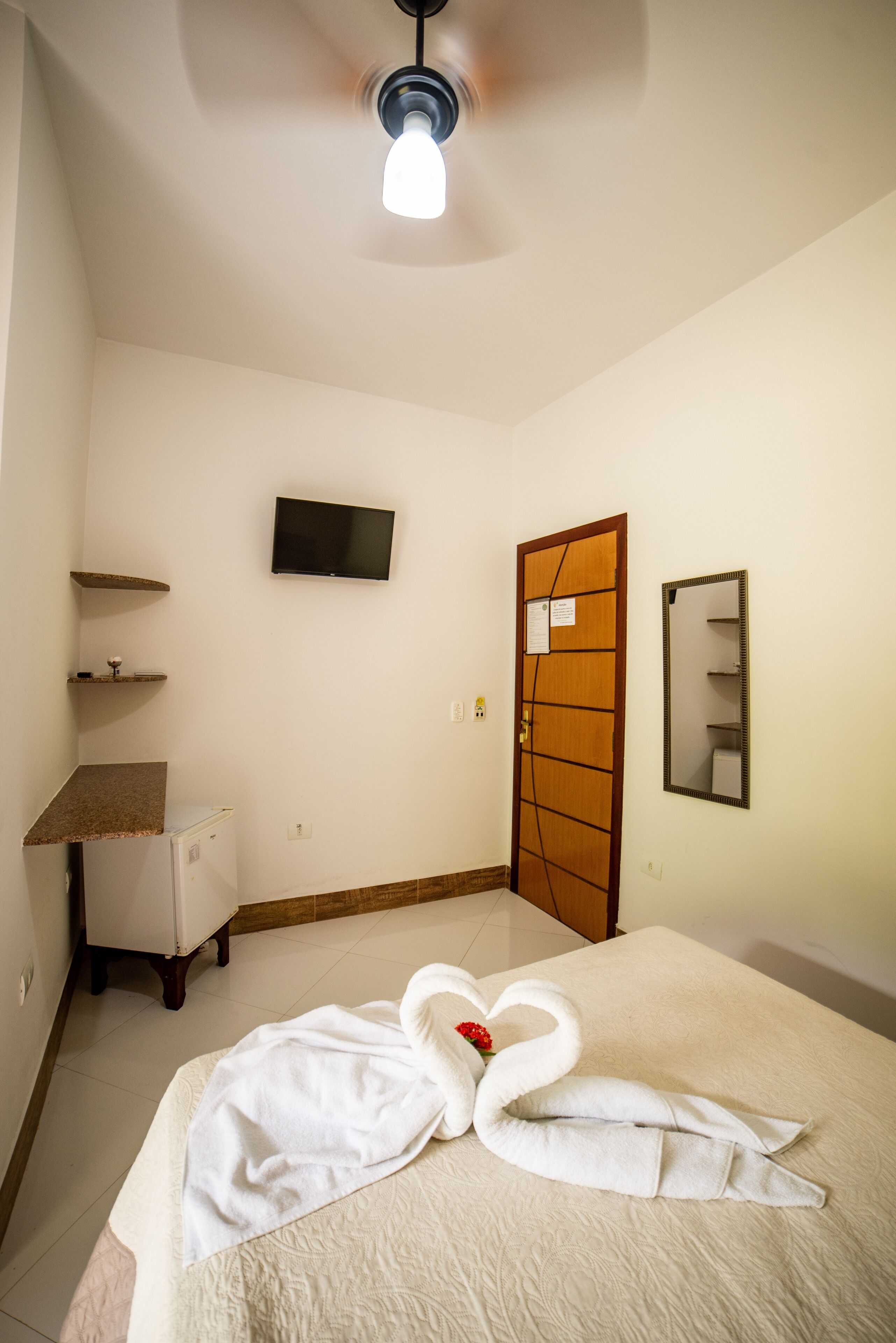 A hotel room with a bed, a television on the wall, a mirror, a door, and a small refrigerator.