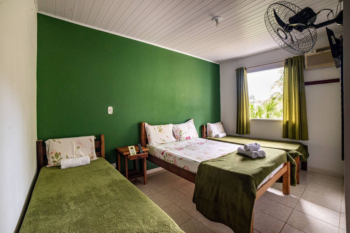 Ein Zimmer mit drei Einzelbetten, jedes mit einer grünen Decke und floralem Bettbezug. Ein kleiner Nachttisch mit Lampe und Fernbedienung ist vorhanden.
