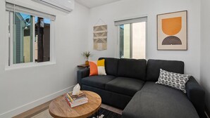 A modern living room with a dark grey sofa, a round wooden coffee table with books, and a wall-mounted air conditioner. - Modern Retreat Steps to the Beach! (San Diego)