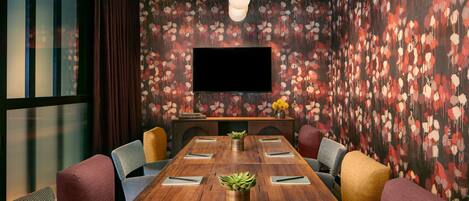 A conference room with a long wooden table, multiple chairs, a flat-screen TV, and a floral wallpaper.