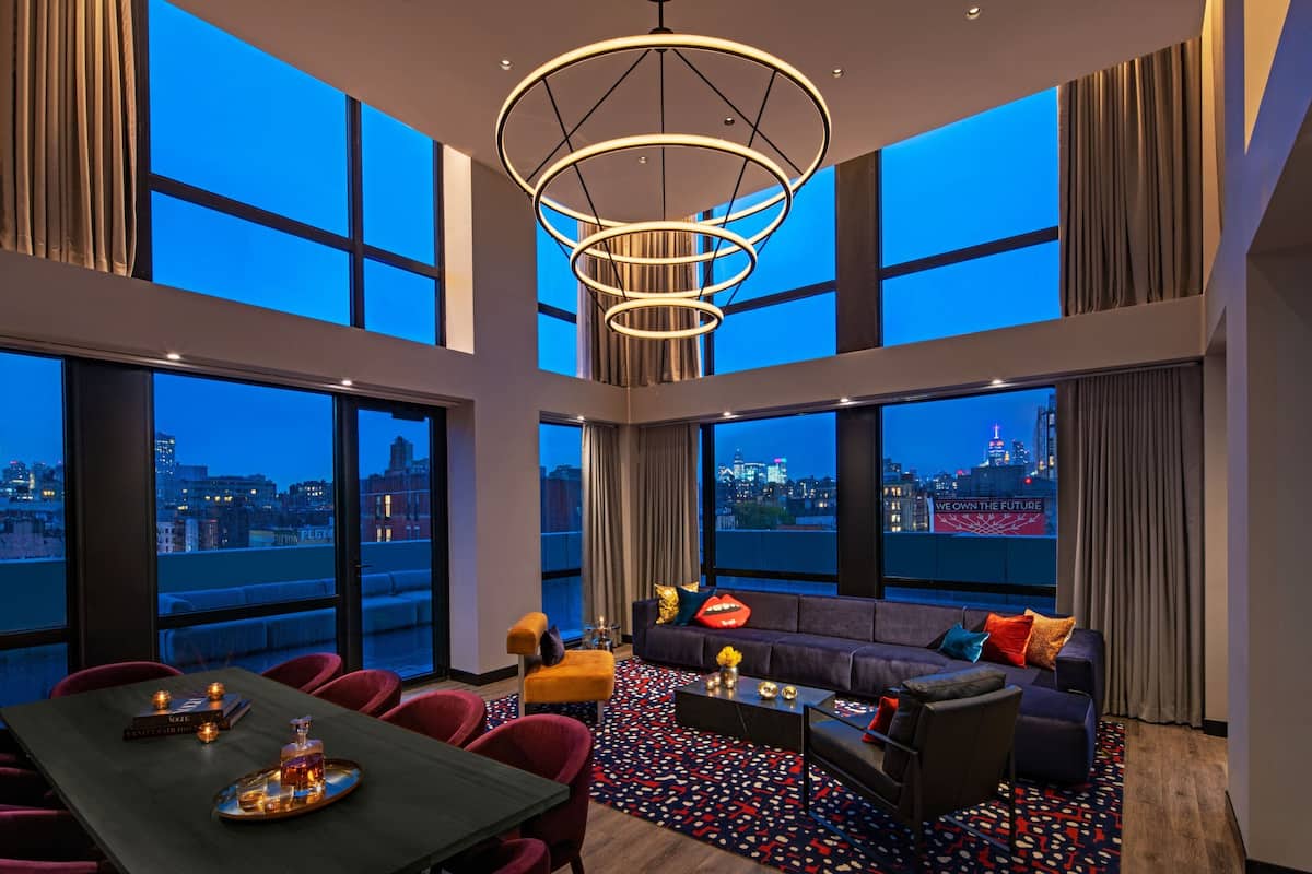 Ein geräumiges Wohnzimmer mit einem modernen Kronleuchter, großen Fenstern mit Blick auf die Stadt, einem bequemen Sofa und einem Essbereich mit Tisch für zwei Personen.