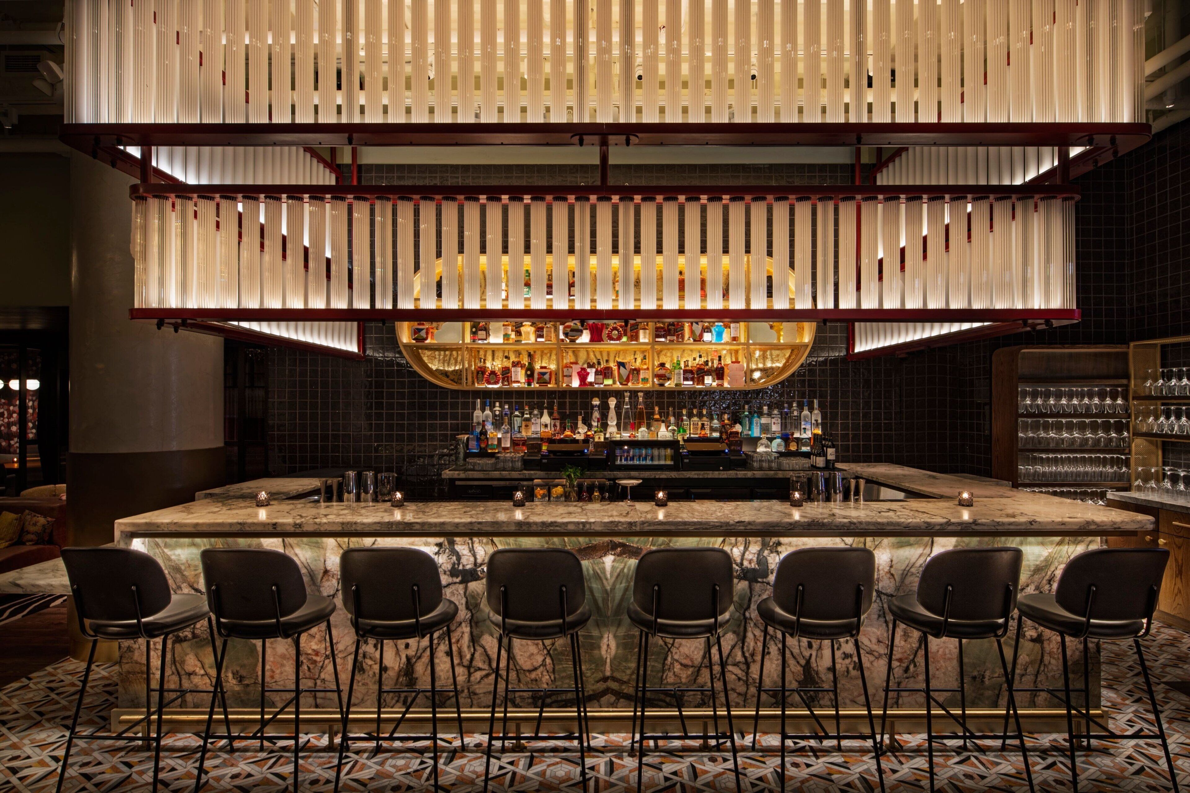 Een bar met een marmeren aanrecht, diverse barkrukken, een scala aan flessen en een bijzondere hanglamp.