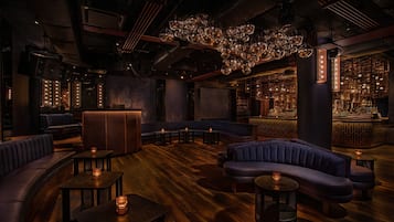 Ett svagt upplyst loungeområde med bekväma sittplatser, små bord och en bar med hyllor fulla av flaskor.