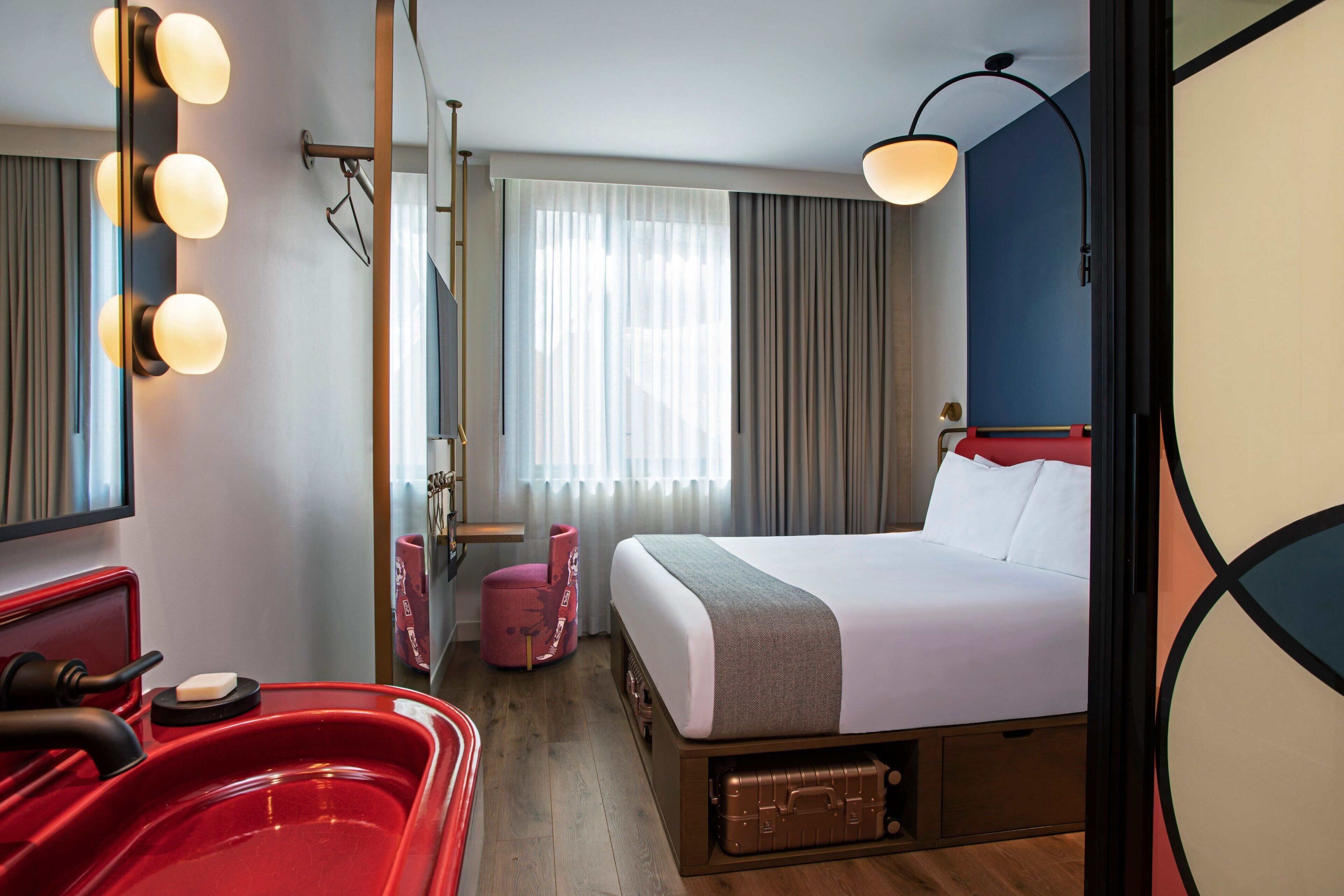 A hotel room with a bed, a red chair, a red sink, a mirror with lights, a window with curtains, and a wall lamp.
