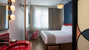 A hotel room with a bed, a red chair, a red sink, a mirror with lights, a window with curtains, and a wall lamp.