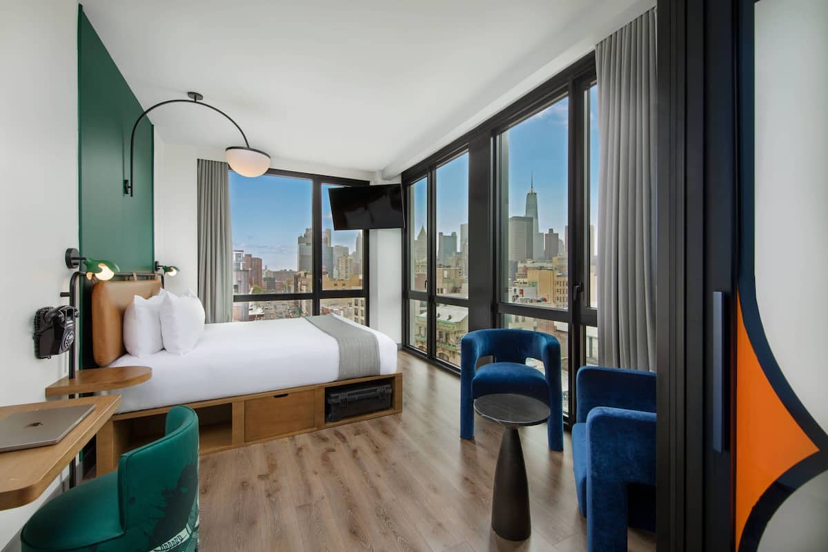 Ein modernes Hotelzimmer mit einem großen Bett, einem Schreibtisch und einem Stuhl. Ein Fernseher ist an der Wand befestigt und es bietet sich ein Blick auf die Stadt durch große Fenster.