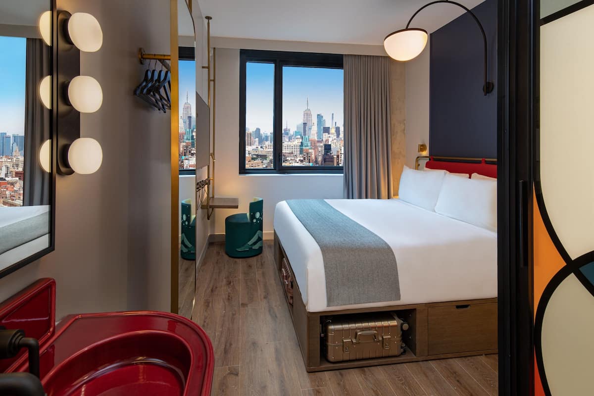 Ein Hotelzimmer mit einem großen Bett, Blick auf die Stadt, einem Schminktisch mit Beleuchtung und einem roten Sessel.