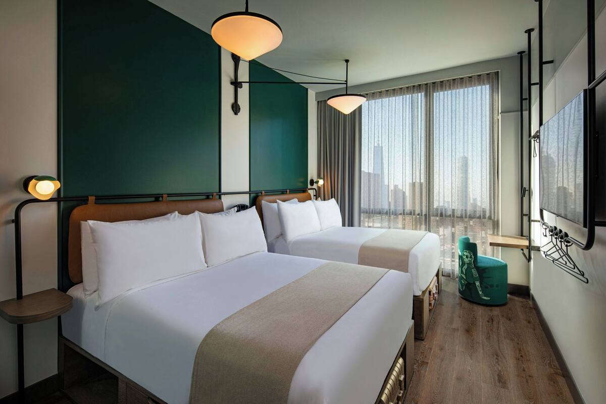 Ein Hotelzimmer mit zwei Betten, Blick auf die Stadt, einem Fernseher und einem grünen Sessel.