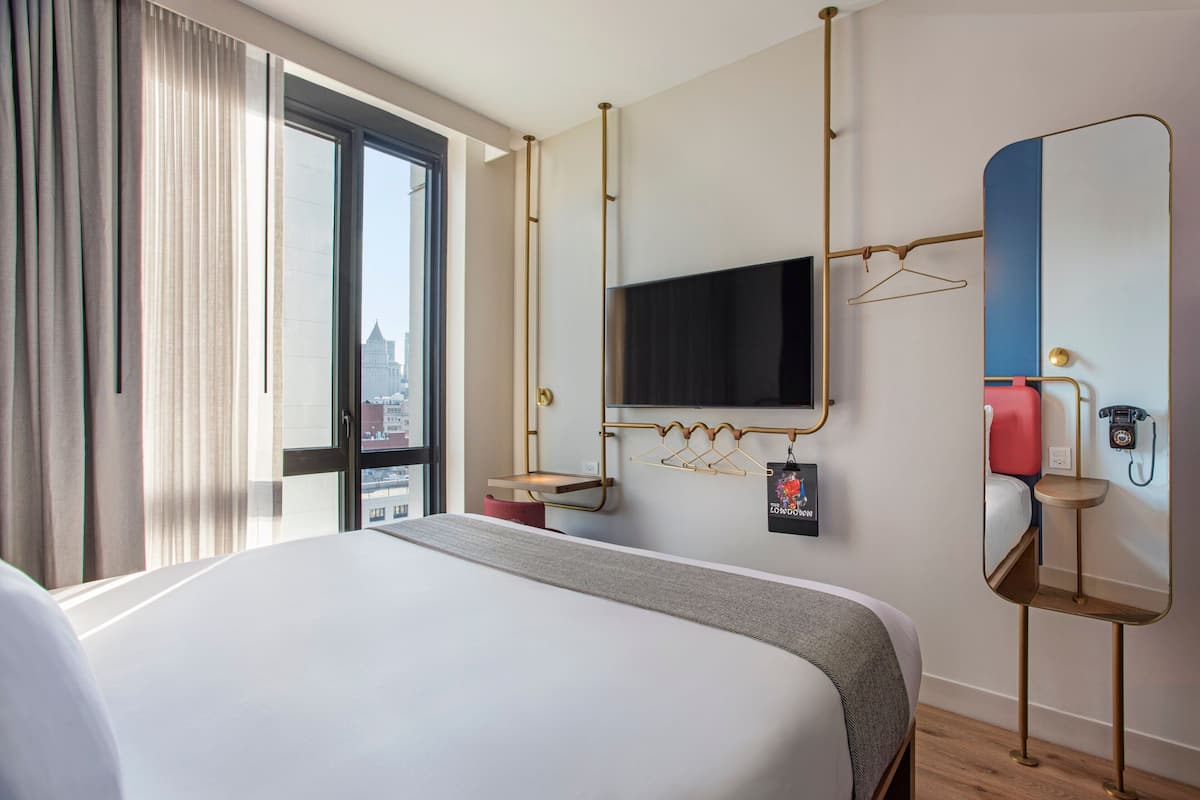Ein Hotelzimmer mit einem großen Bett, einem an der Wand befestigten Fernseher, einem Spiegel und einem Fenster mit Vorhängen.