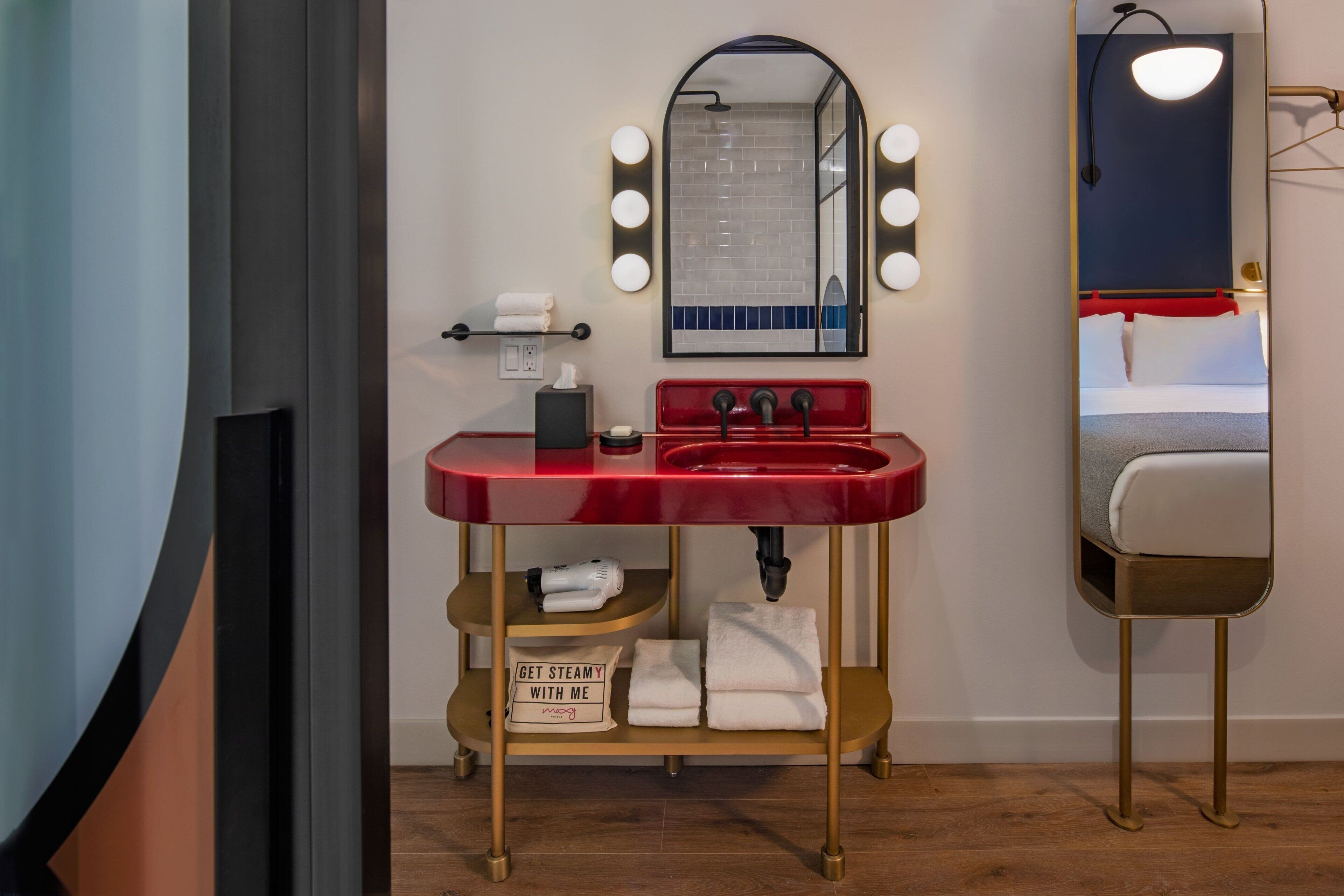 Un baño con lavamanos rojo, espejo de tocador con luces y un perchero para toallas.