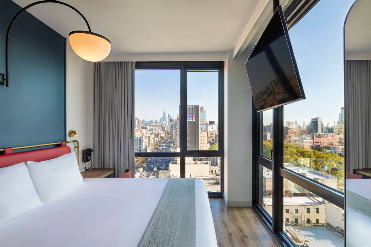 Ein Hotelzimmer mit einem großen Bett, einem an der Wand montierten Fernseher und Blick auf die Stadt durch die Fenster.