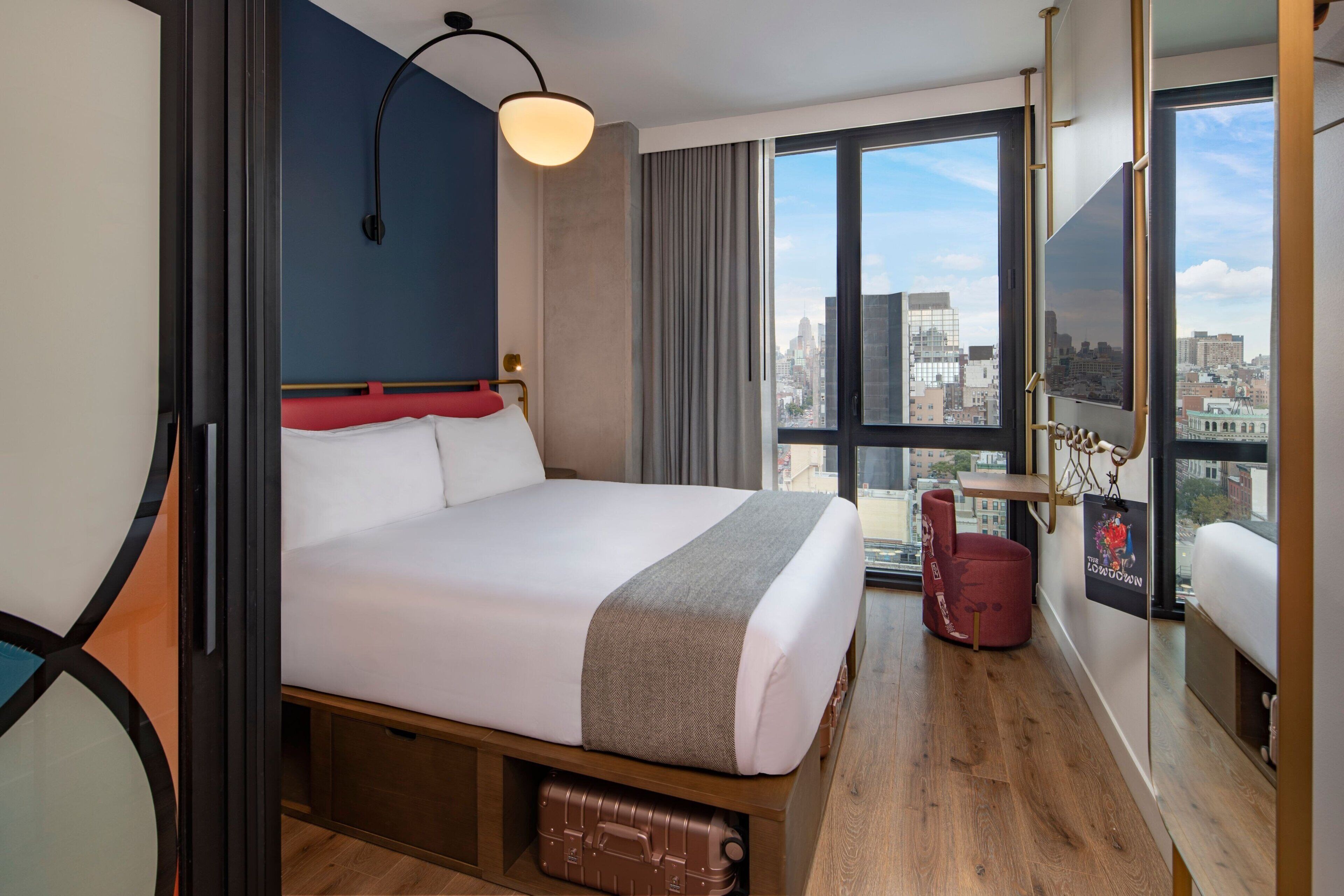A hotel room with a large bed, a city view, a red chair, and a suitcase under the bed.