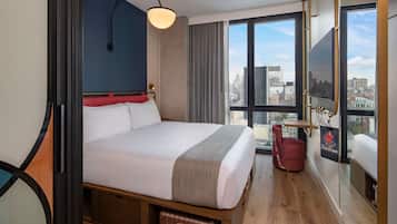 A hotel room with a large bed, a city view, a red chair, and a suitcase under the bed.