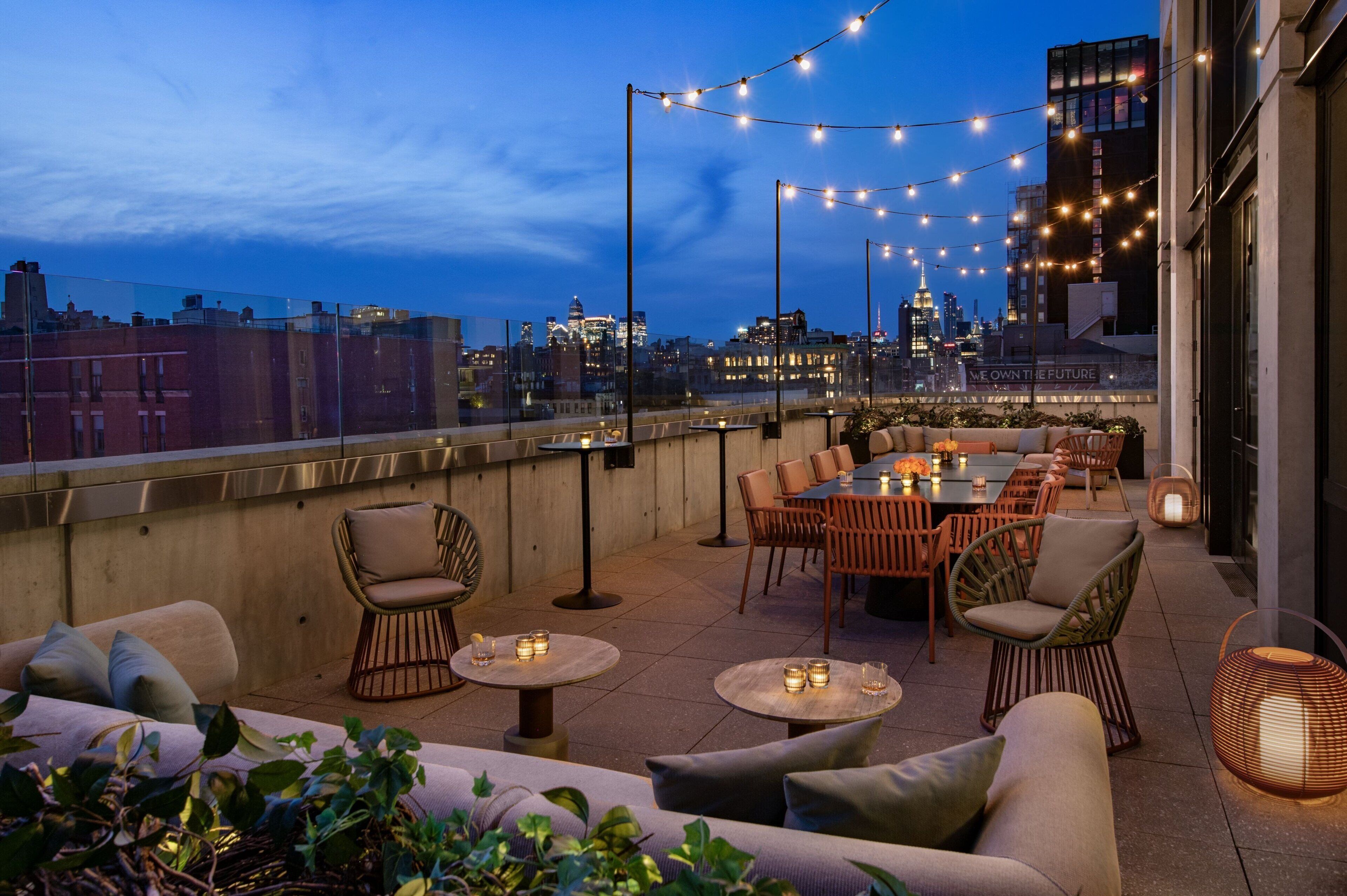 En tagterrasse med udendørs møbler, lyskæder og udsigt til byens skyline.